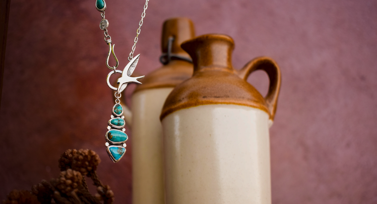 A silver-toned necklace with a hummingbird pendant and a vertical drop of turquoise stones, displayed in front of two rustic ceramic pitchers against a textured mauve background.
