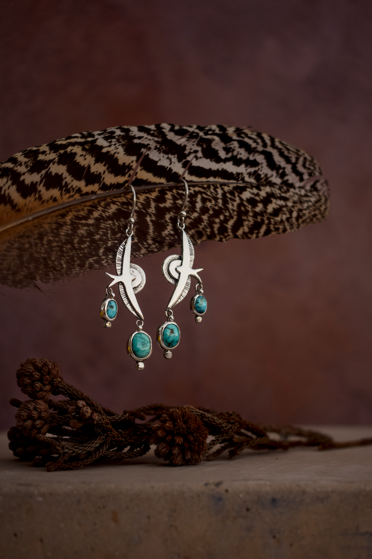 Silver dangle earrings with turquoise blue stone drops, hanging from a large brown patterned feather against a dark brown background.