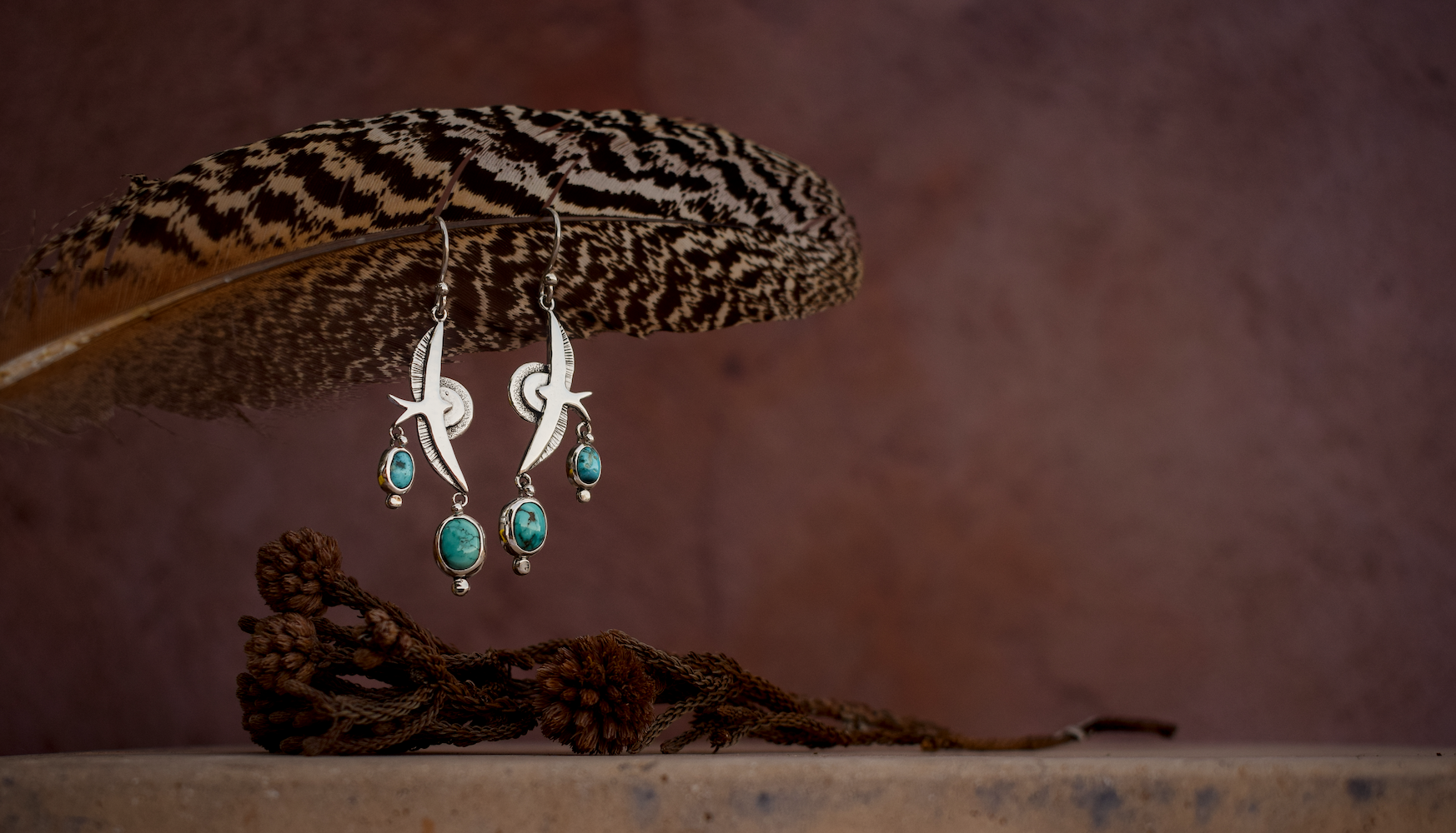 A pair of silver dangle earrings with turquoise blue beads, hanging from a large brown and black patterned feather against a textured brown background.